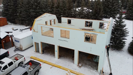 An in-progress image of a residential home being built with Nudura ICF. The barn-style home is surrounded by snow, pine trees, and pick-up trucks.
